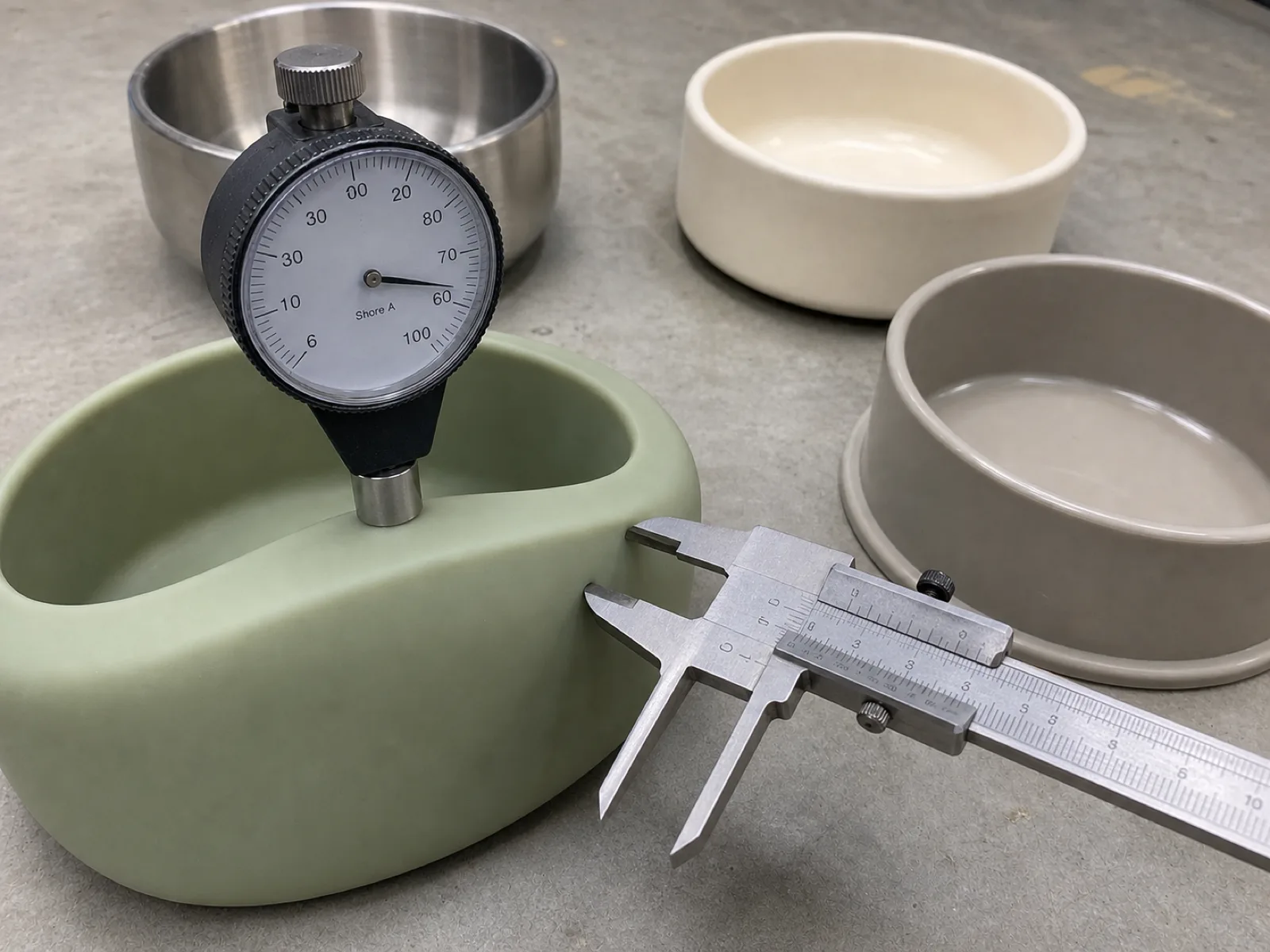Durometer and material comparison of a food grade silicone pet bowl on a QC bench — a durometer pressed into the rim of a sage platinum-cured pet bowl reading in the low sixties, staged beside a grade-304 stainless steel bowl, a ceramic bowl, and a rigid plastic bowl for material comparison, with a caliper measuring wall thickness on a neutral concrete workshop floor.