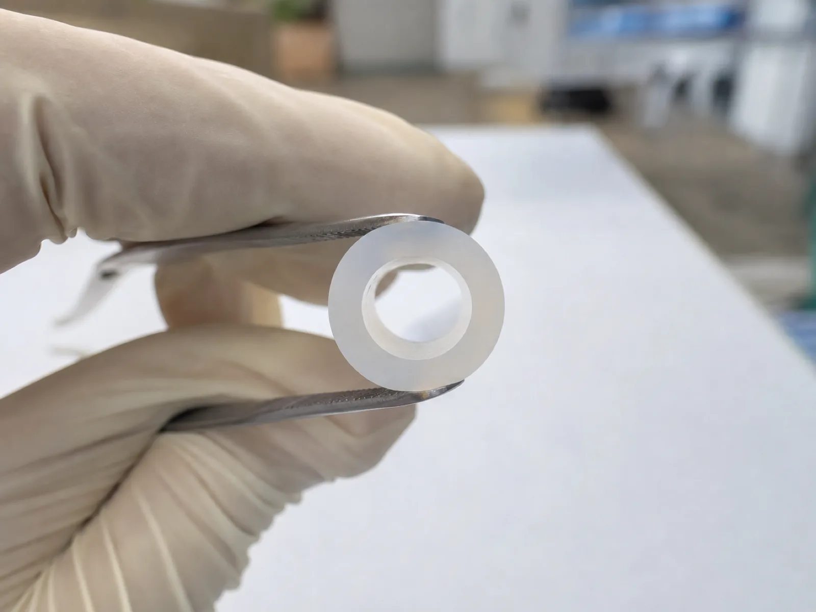 Close-up cross-section of a food grade silicone tubing coil freshly cut with a razor, showing the concentric inner and outer diameter and the smooth translucent wall, held with tweezers over a matte white QC background