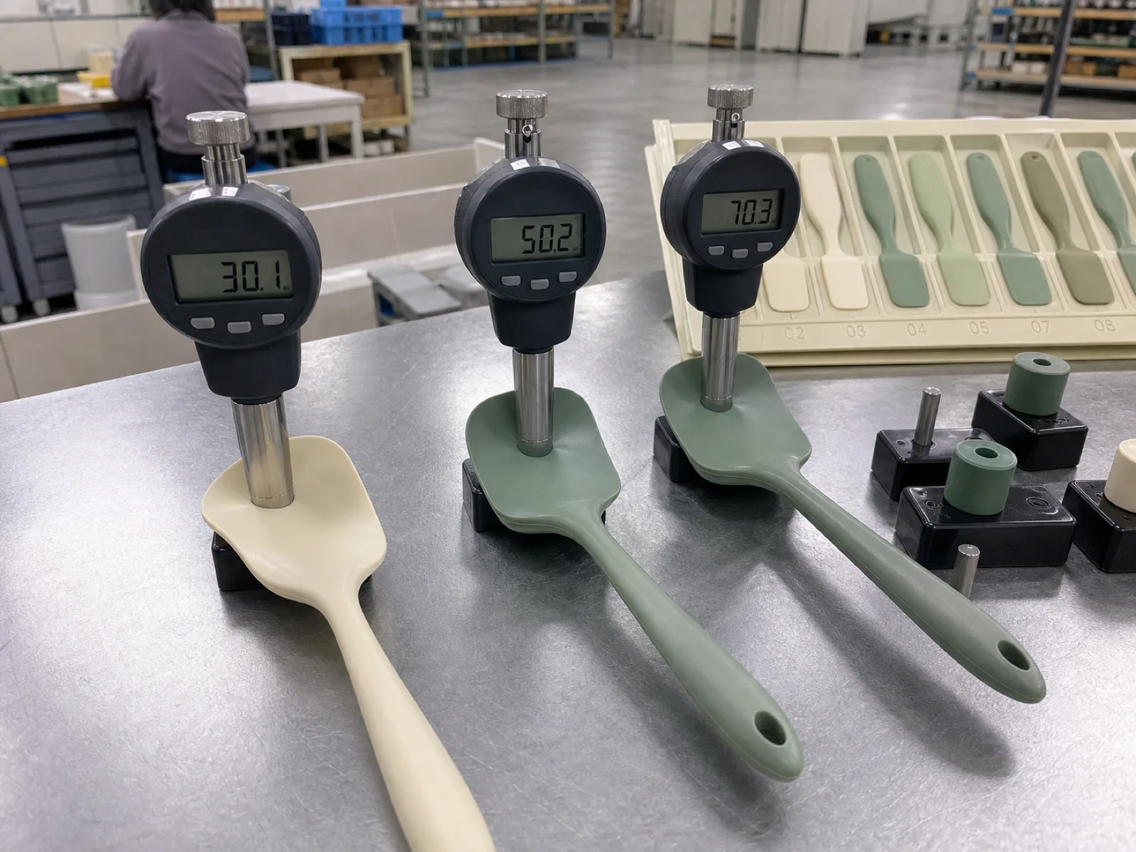 Three cream and sage silicone spatula heads of different hardness on a QC bench, each being flexed against a digital Shore A durometer reading 58, 65, and 71, calibration blocks and a cavity-numbered sample tray in the muted-palette background.