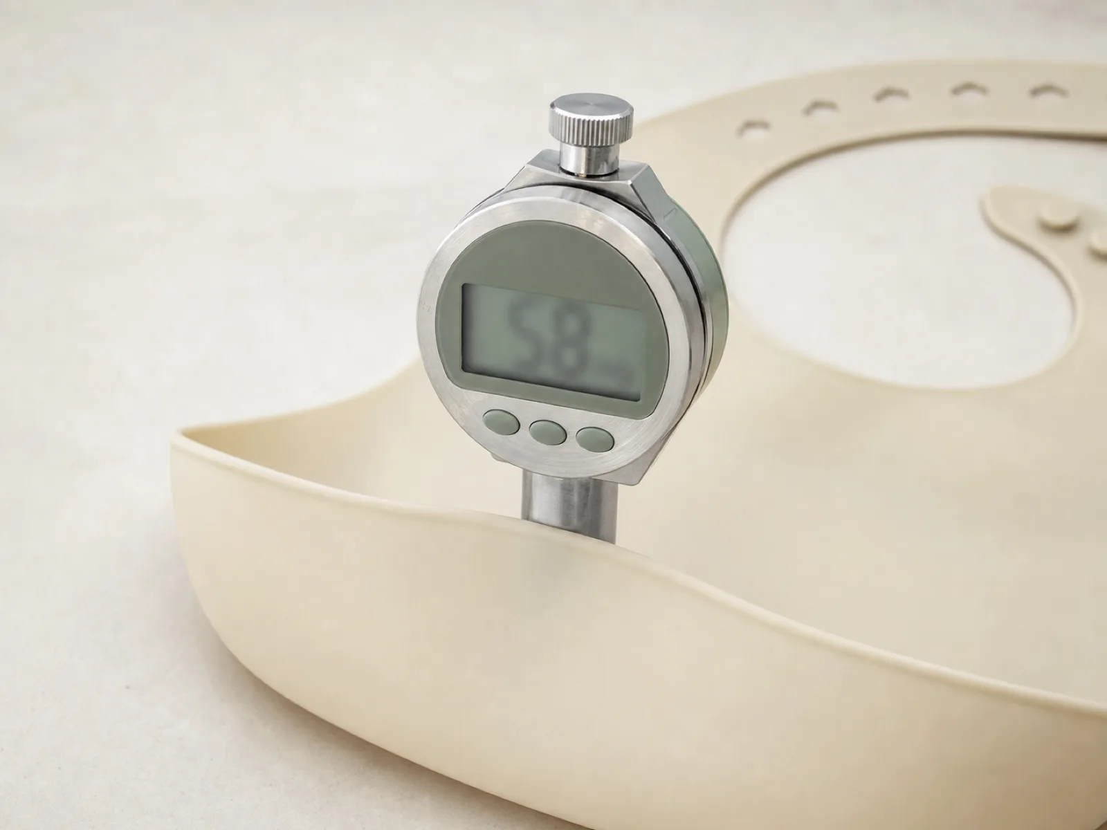Shore A durometer measurement on the catch-pocket rim of a platinum-cured silicone baby bib at the Wetop QC bench — 55 Shore A target with 3 mm indentation depth, D65 light-box background.