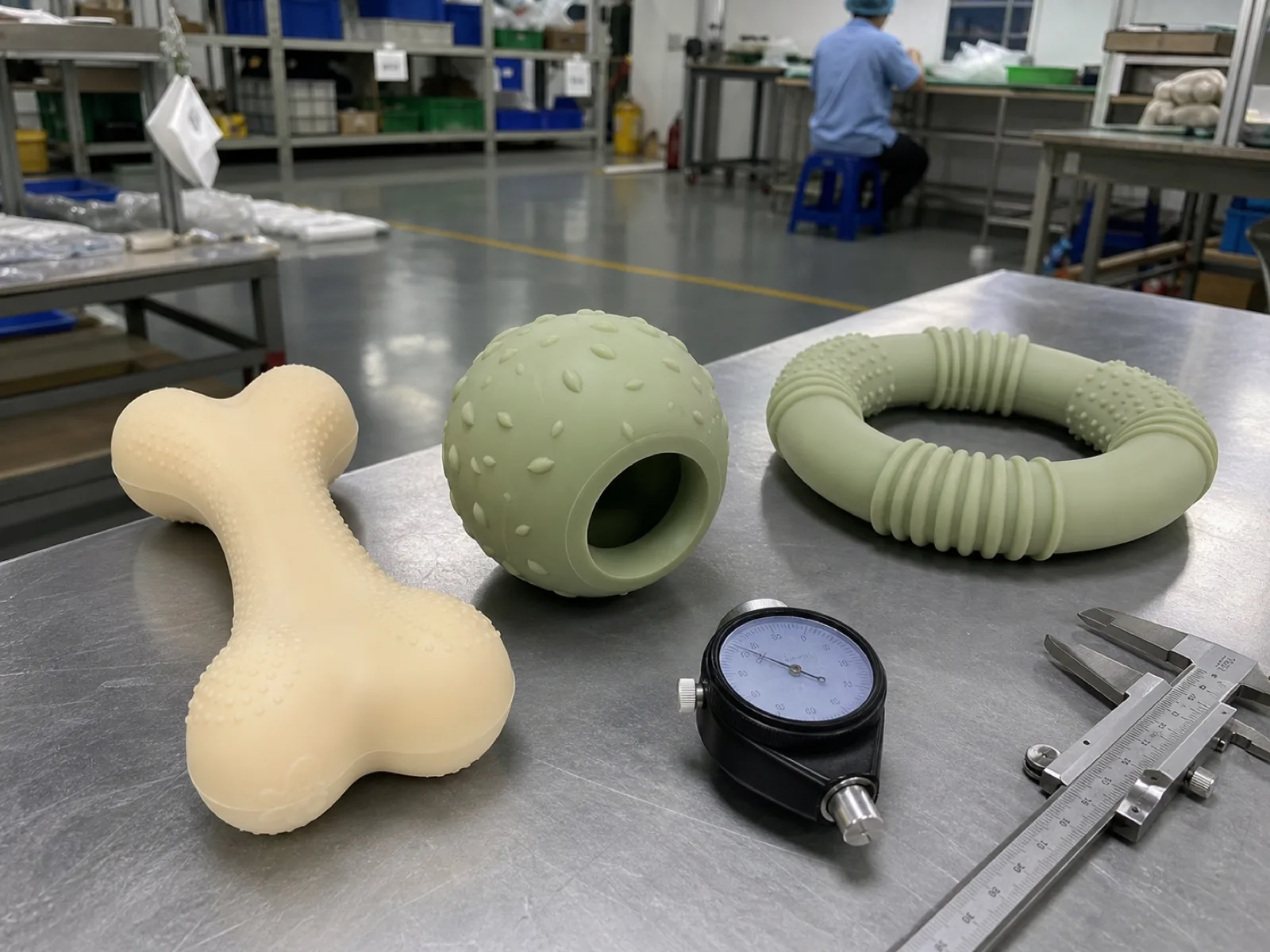 Anonymized platinum-cured silicone dog toys — a chew bone, a treat-dispensing ball, and a tug ring in cream and sage — arranged on a QC bench beside a durometer gauge and calipers at a Dongguan silicone factory.