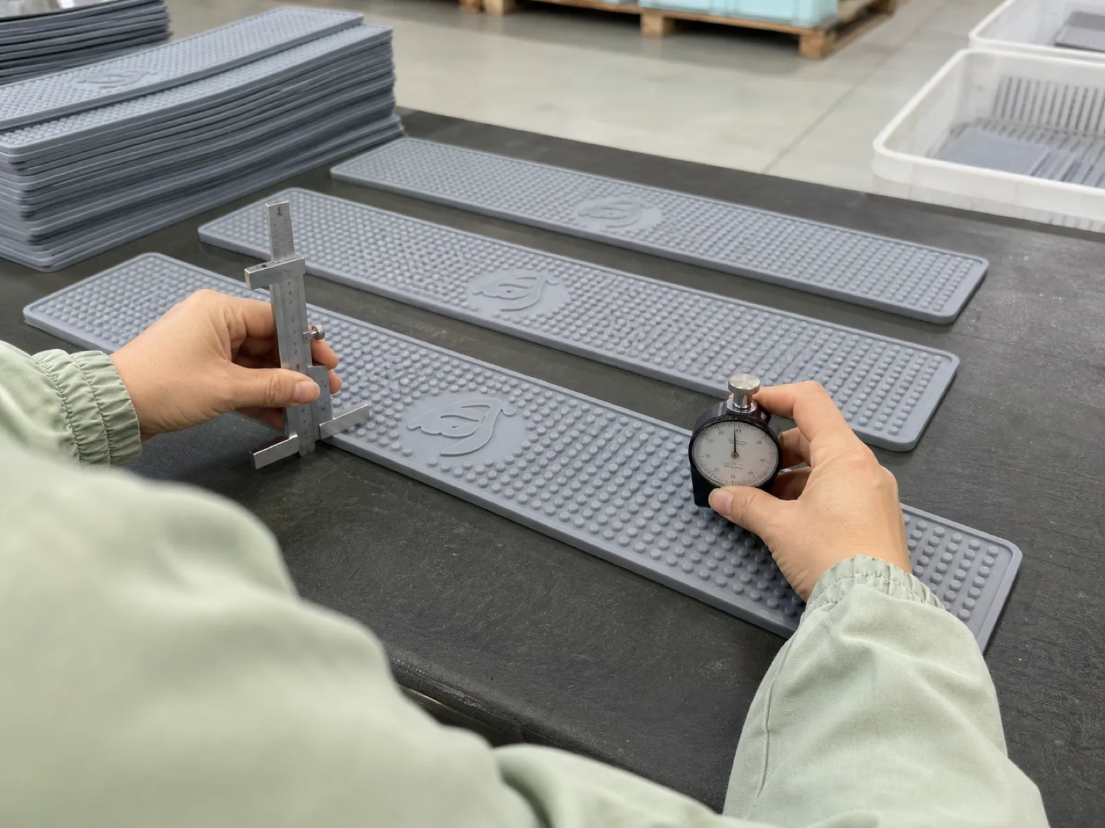QC bench inspection on a bulk hospitality silicone drip mat program at Wetop's Dongguan floor — technician measuring debossed logo depth and Shore A hardness on 600 mm bar-rail drip mats under D65 workshop lighting, anonymized restaurant-service samples