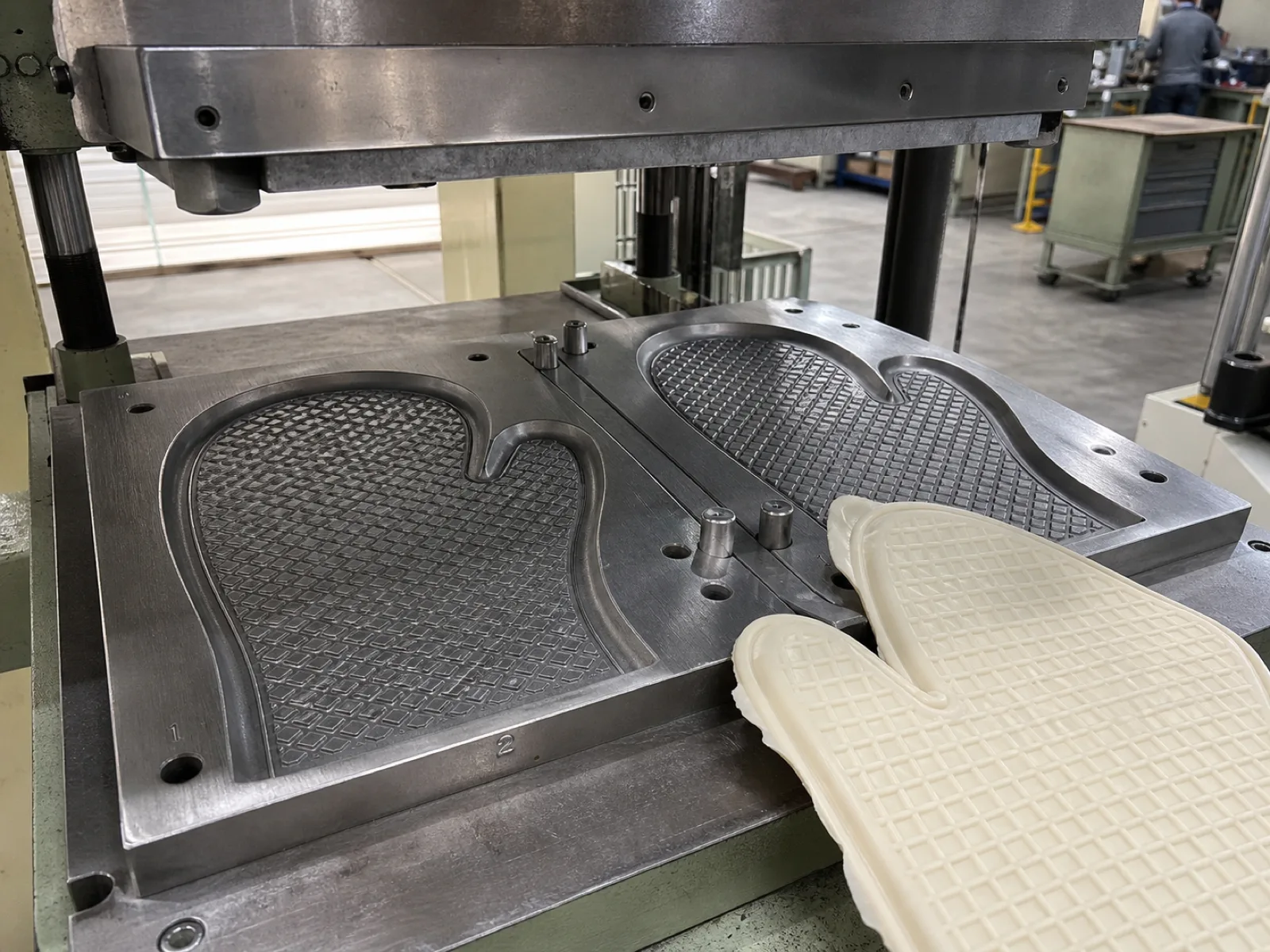 Silicone oven mitt compression mold on a Wetop press bed — a two-cavity mold with the criss-cross grip texture engraved into each cavity face, cavity ID numbers etched at the edge for QC traceability, cream silicone flash visible around a demolded shell, hydraulic press platen above.