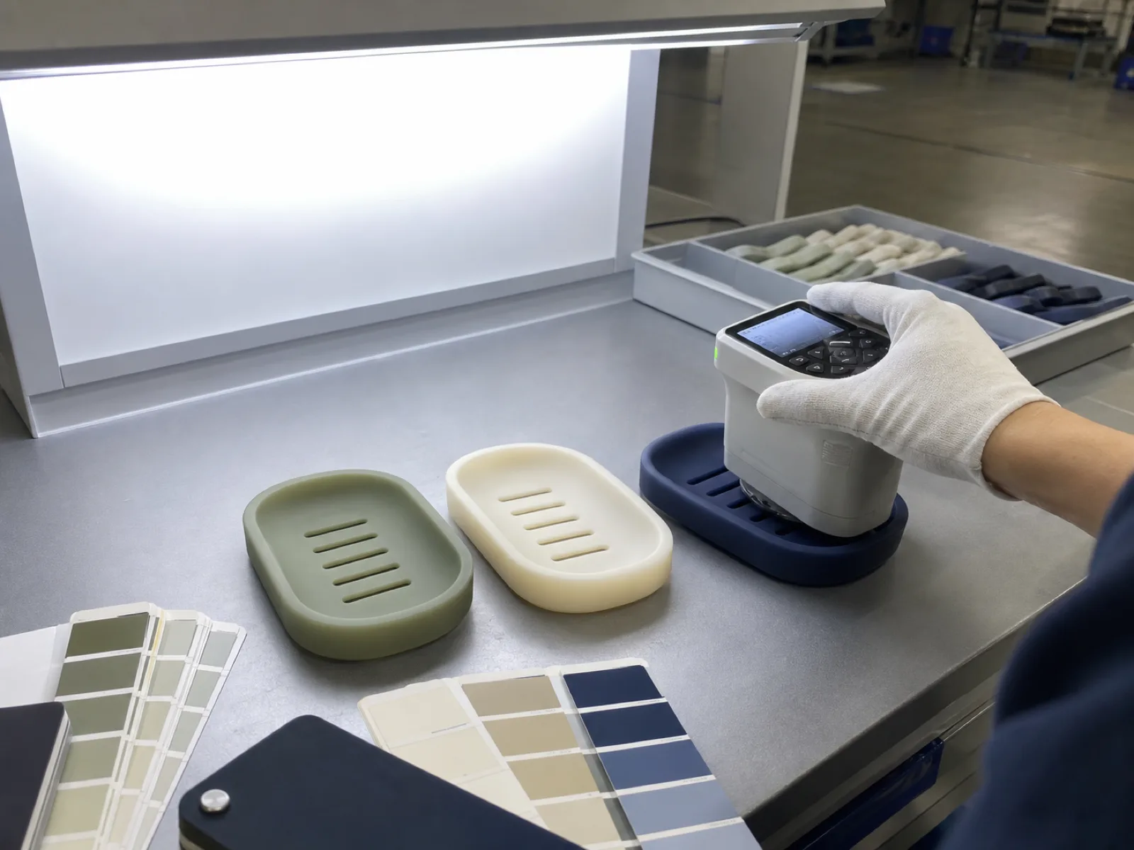 Wetop Silicone Pantone color verification bench with silicone soap dish samples in muted sage, warm off-white, and matte navy under D65 daylight-simulation light-box with a colorimeter measuring delta E readings on a Wetop QC bench in Dongguan