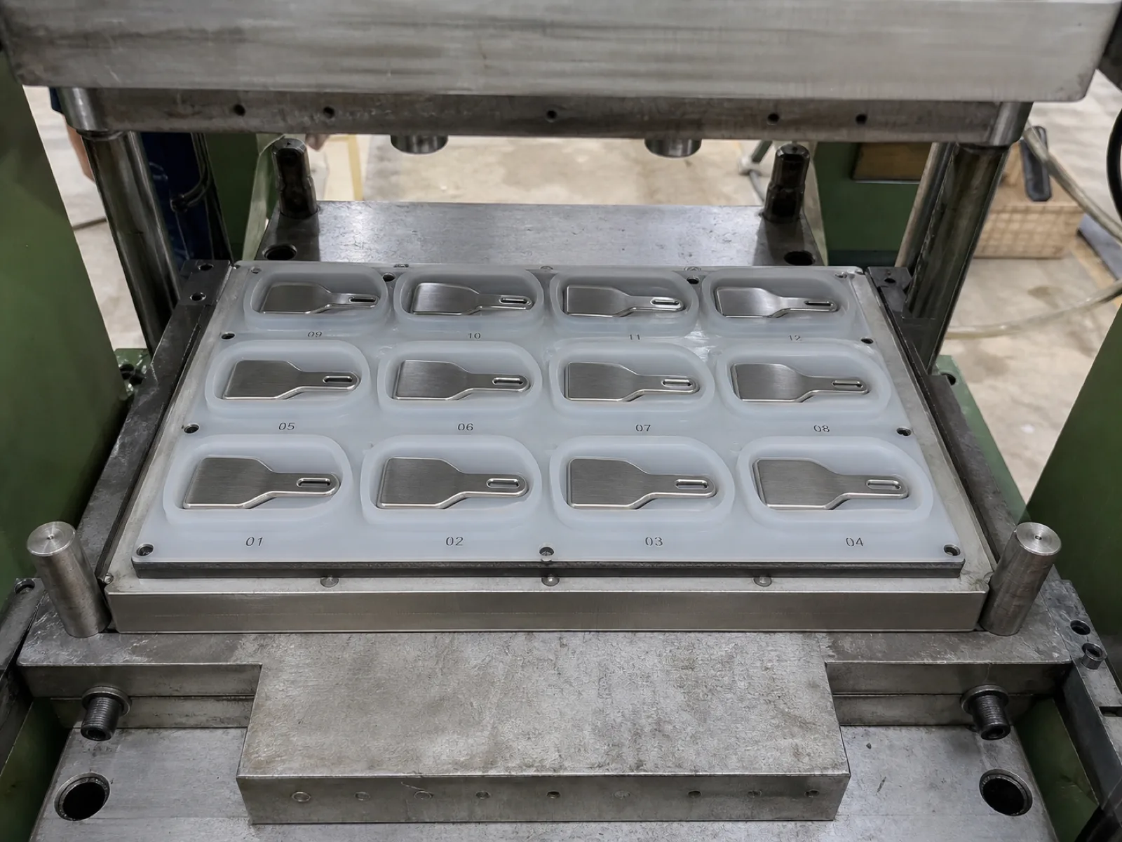 Multi-cavity silicone spatula mold on a Wetop compression press bed — twelve cavities laid out in a 3×4 grid, stainless steel core inserts pre-loaded in each cavity, cavity numbers etched at the edge for QC traceability, hydraulic press platen visible above.