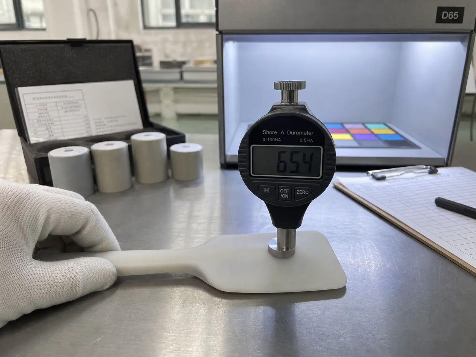 Silicone spatula QC inspection bench at Wetop — a Shore A digital durometer reading 65.4 on a freshly demolded spatula head, engineer's gloved hand steadying the sample, calibration blocks and D65 light box color-check station visible in the background.