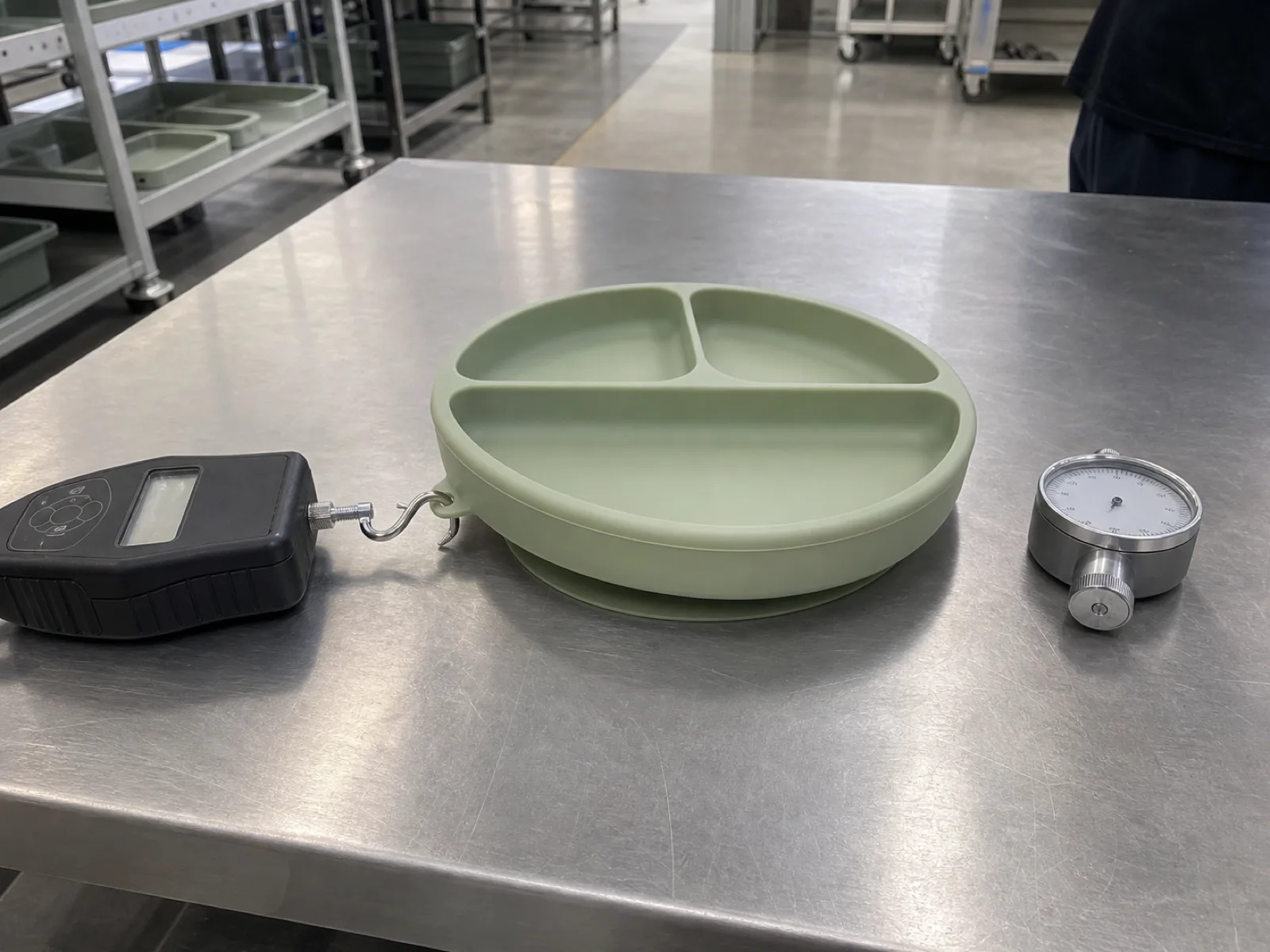 One-piece platinum-cured silicone suction placemat plate in sage-green on a QC bench with pull-force gauge and durometer, Dongguan silicone factory
