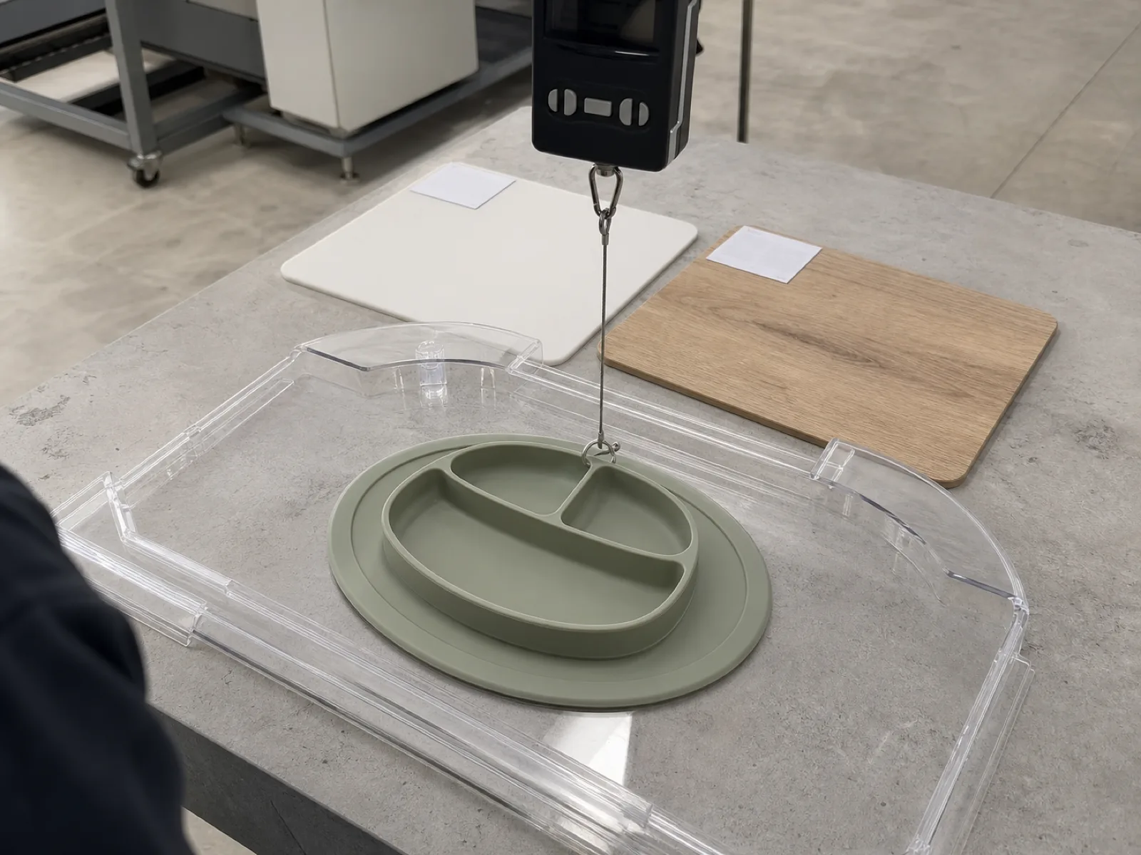 Digital pull-force gauge measuring straight-up suction release on a sage silicone suction placemat plate mounted to a smooth acrylic high-chair tray sample, QC bench