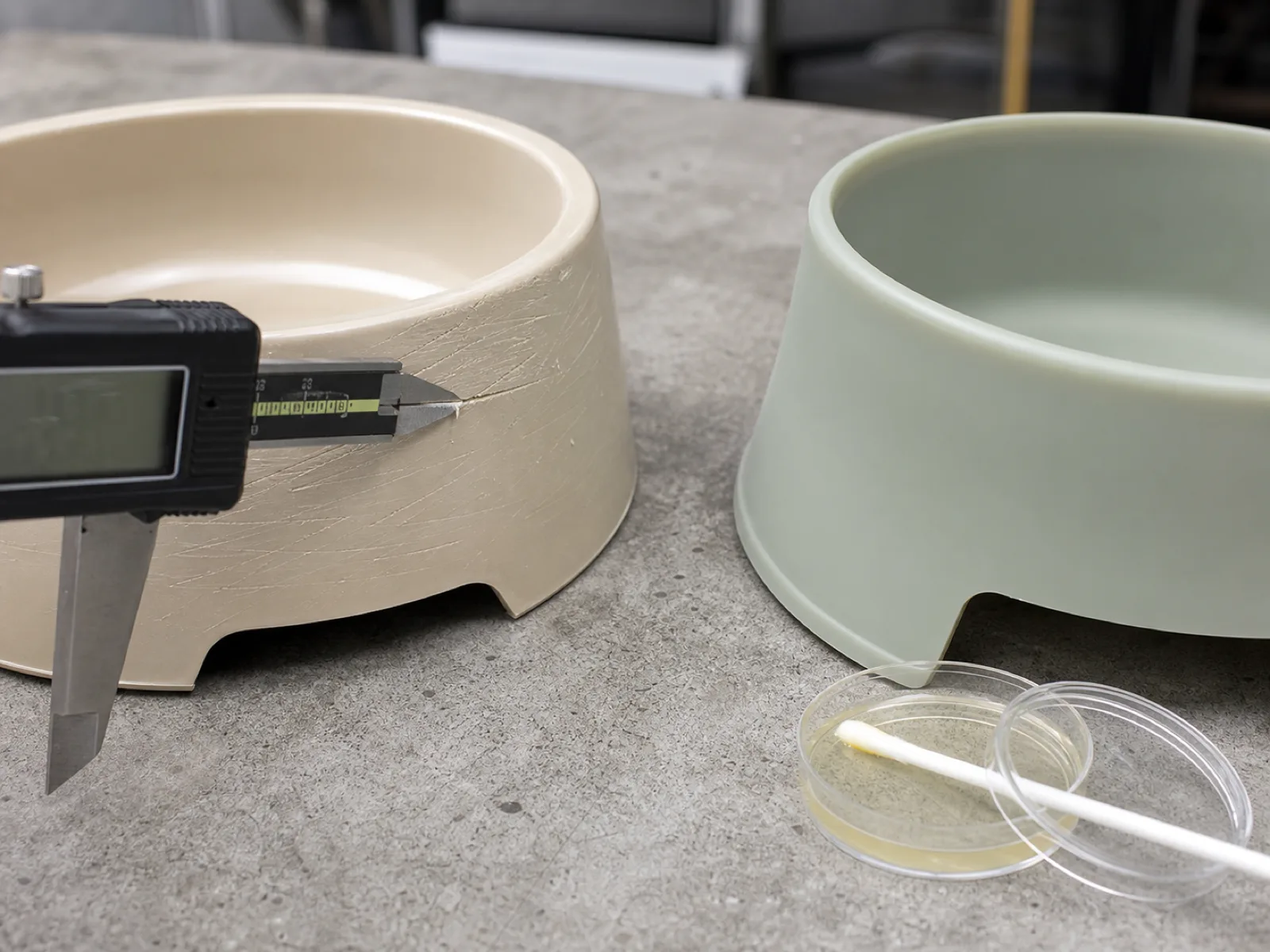 QC bench close-up comparing a scored plastic dog bowl surface against a non-porous food-grade silicone bowl wall, with a digital caliper measuring scratch depth and a swab plate staged under D65 workshop light on a neutral concrete bench.