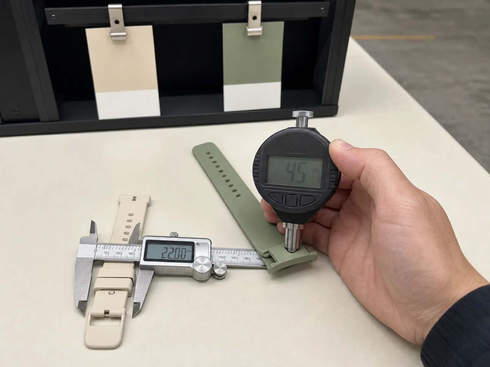QC bench inspection of silicone watch bands at Wetop — technician back-of-hand only, digital durometer showing Shore A 45 reading pressed against a sage strap, calipers on a 22 mm lug width, D65 light box behind for Pantone TCX color-drift check, cream backdrop and matte concrete floor