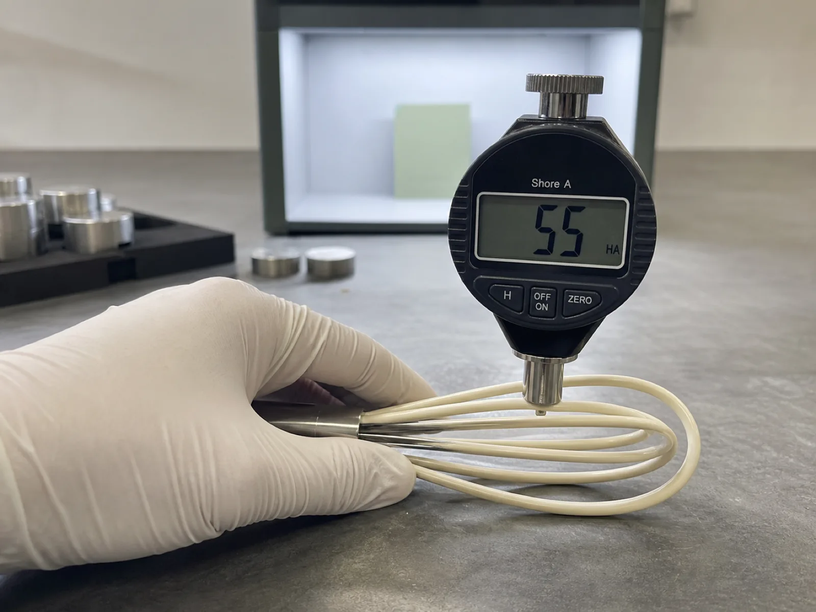 Silicone whisk QC inspection bench at Wetop — a Shore A digital durometer pressed against the coating of a freshly demolded balloon whisk loop reading 55.2, engineer's gloved hand steadying the sample, calibration blocks and a D65 light box color-check station in the background.