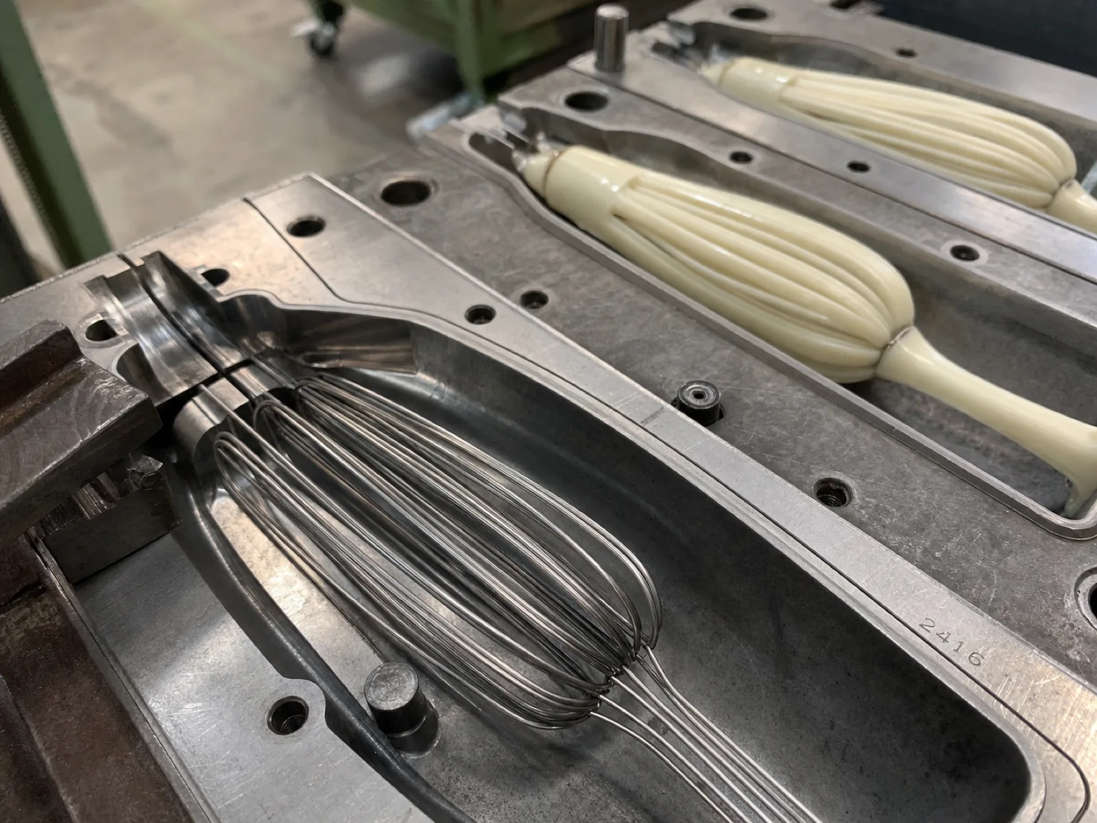 Close-up of a silicone whisk over-mold cavity on a Wetop compression press — a bundle of primer-treated stainless steel wire loops seated in the mold before the silicone shot, cream-colored coating already flowed around adjacent loops, cavity edge etched with a QC number under D65 workshop light.